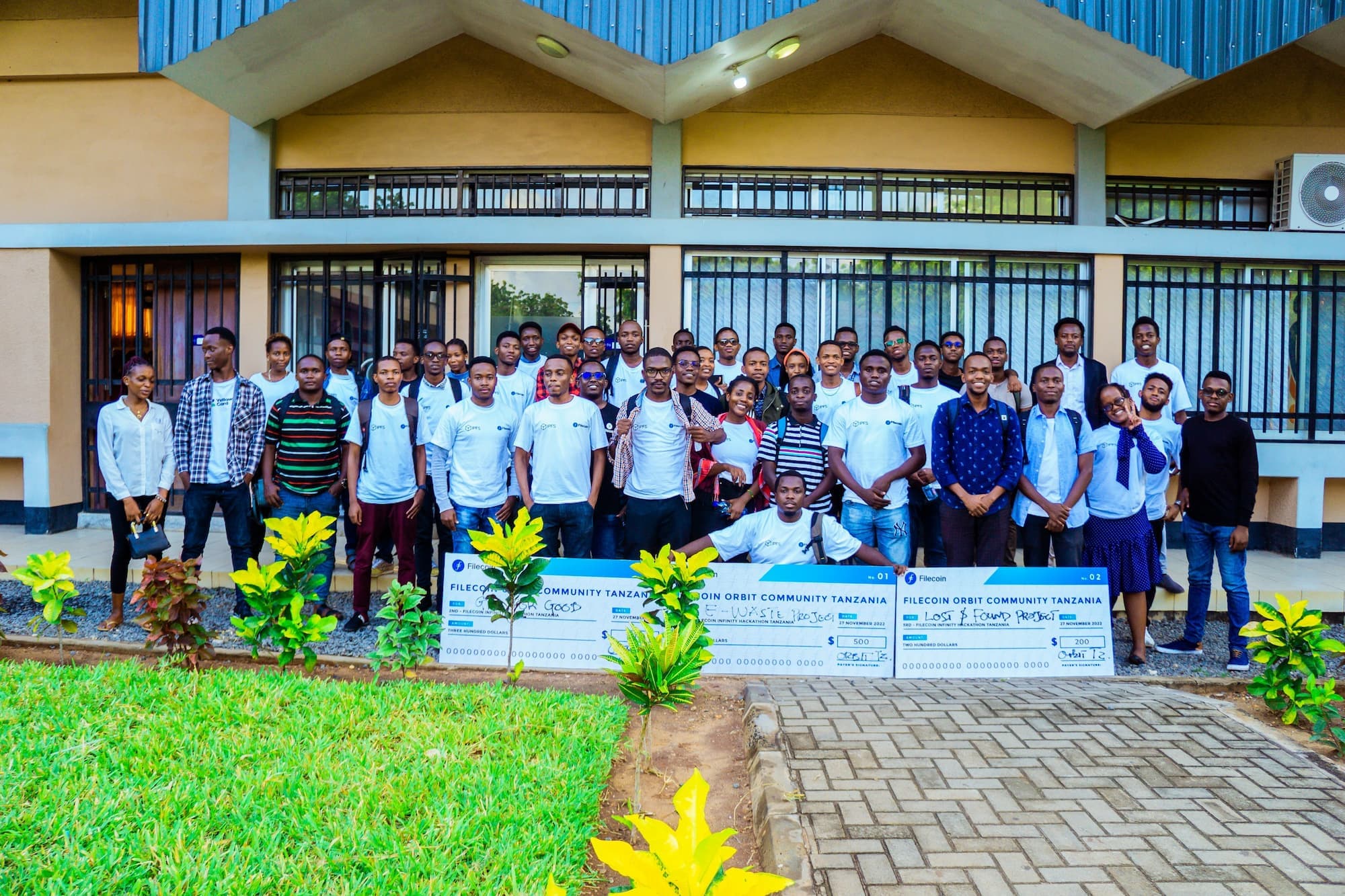 A large group of people posing for a group photo outside a building at the Filecoin Orbit Community event in Tanzania. Many of the attendees are wearing white Filecoin t-shirts. Some individuals in the front row are holding large checks as awards for various projects. The group is standing in front of a building with large windows and plants in the foreground.